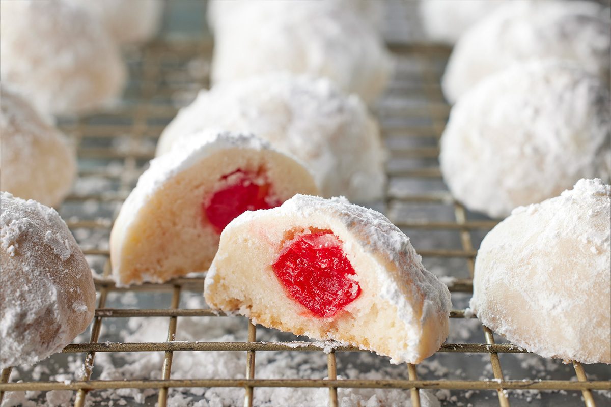 Closeup shot of cherry snowball cookies dusted with powdered sugar on a cooling rack, with one broken open to reveal the vibrant red cherry filling inside;