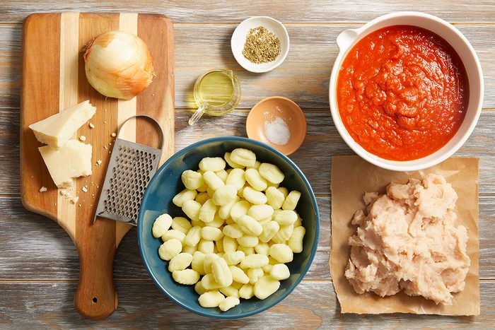 Ingredients for a meal arranged on a table: gnocchi in a bowl, ground meat on paper, tomato sauce in a bowl, an onion, parmesan cheese, a grater, olive oil, salt, and dried herbs.