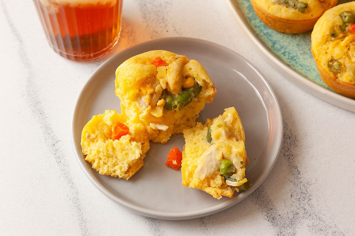 Close shot of a plate with a Chicken Potpie Muffin cut in half, revealing chicken and vegetables, with iced tea and more muffins on a blue plate in the background;