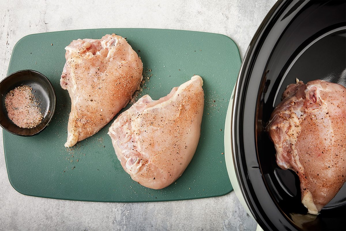 Raw, seasoned chicken breasts on a green cutting board next to a small bowl of seasoning, with one chicken breast placed inside a black slow cooker on a gray surface.