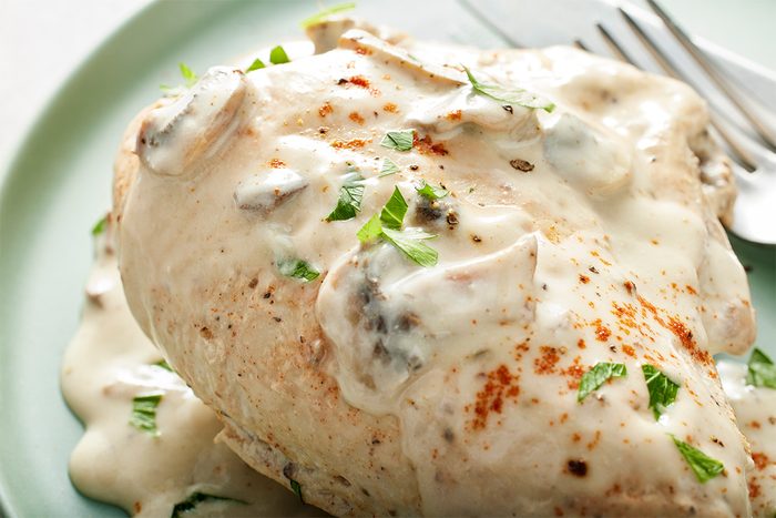 A cooked chicken breast topped with creamy white mushroom sauce, garnished with chopped parsley and a sprinkle of paprika, served on a light green plate with a knife and fork beside it.