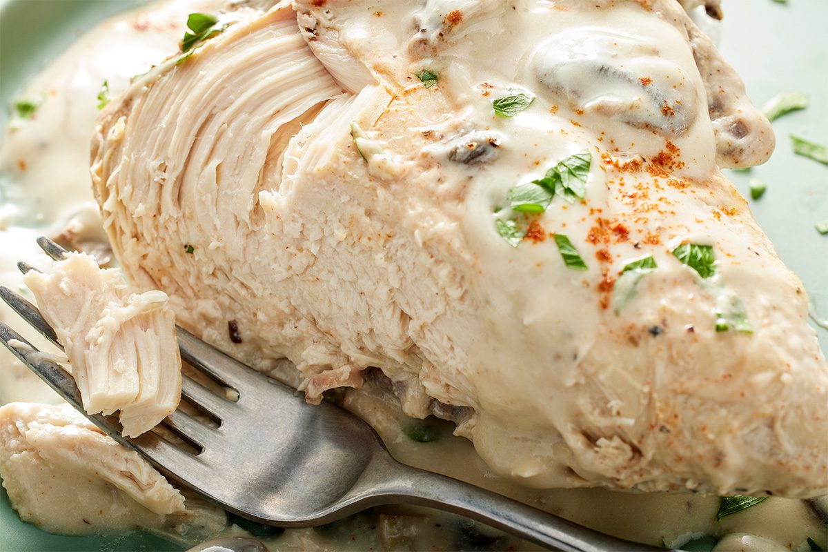 A close-up of a cooked chicken breast covered in creamy sauce, garnished with chopped herbs, with a fork holding a bite-sized piece in the foreground.