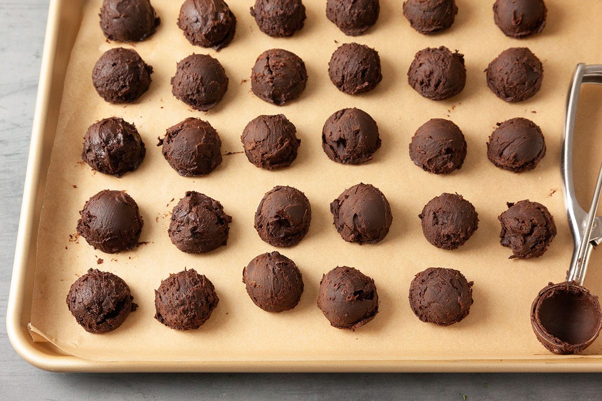 3/4th shot of a baking sheet lined with parchment paper shows rows of chocolate cookie dough balls and a metal cookie scoop with one spot empty