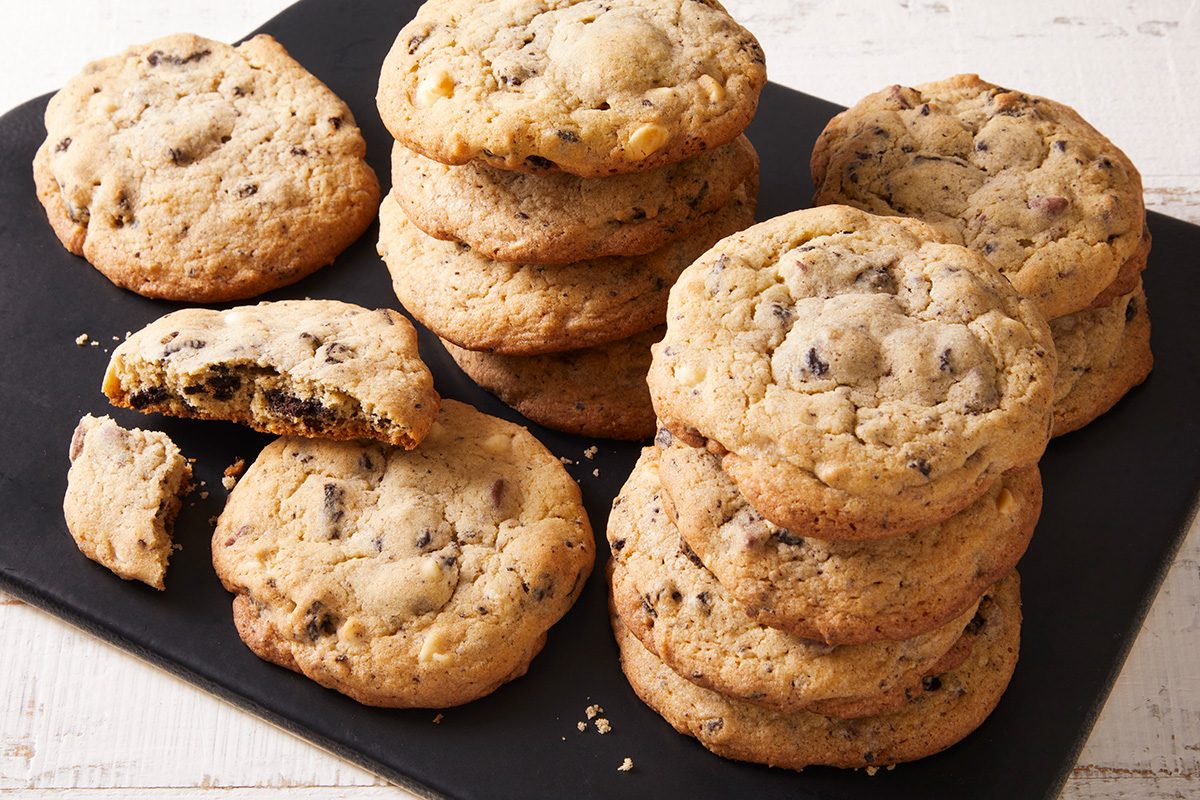 A pile of chocolate chip cookies is arranged on a black slate board. Some cookies are stacked, while others are lying flat. One cookie is broken in half, showing its chocolate chip-filled center.