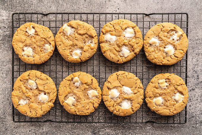 Eight golden brown cookies with white marshmallow pieces are arranged in two rows on a black wire cooling rack, set on a gray textured surface.