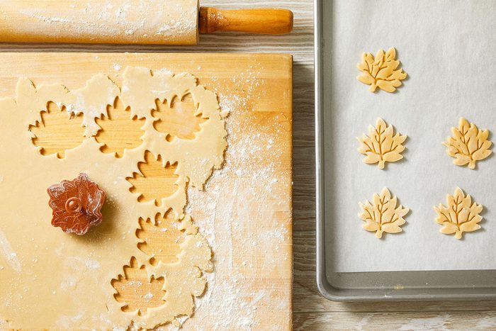 Overhead shot of a wooden rolling pin and board hold leaf-shaped cookie dough cutouts and a cookie cutter while five cookies rest on a tray dusted with flour