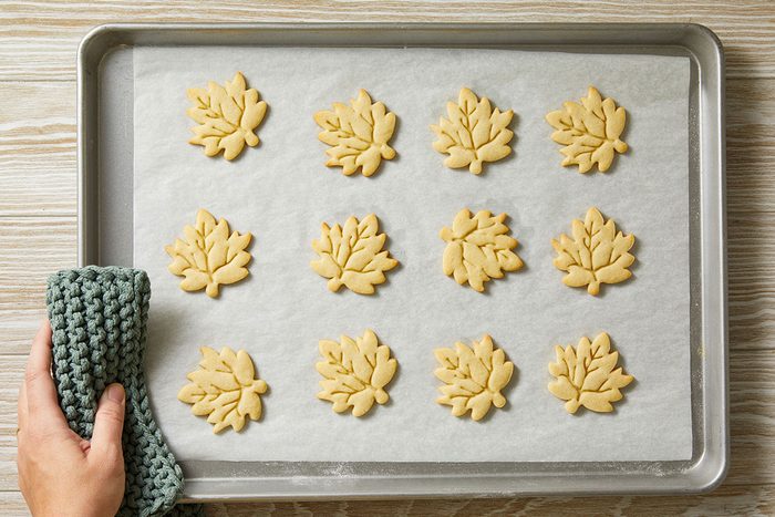 Overhead shot of a hand with a textured oven mitt lifts a tray of twelve unbaked leaf-shaped cookies They are lined up on parchment over wood