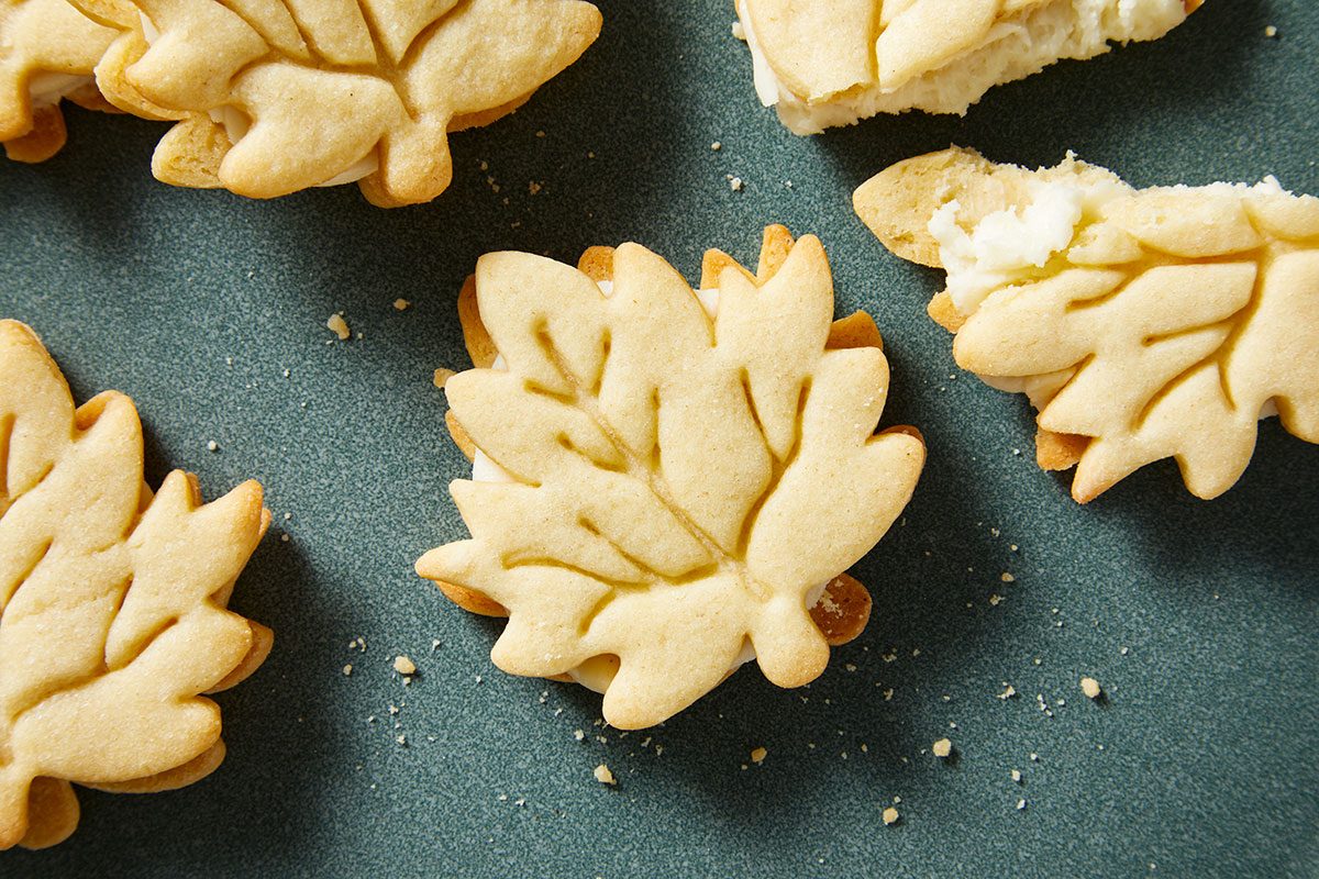Horizontal Shot of Copycat Trader Joe's Maple Leaf Cookies are stacked on a dark plate and creamy filling peeks out more cookies rest on a light wood surface