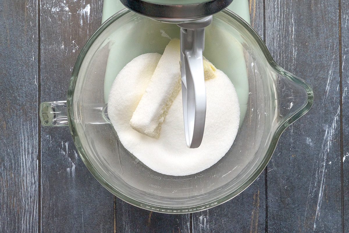 Overhead shot of a glass mixing bowl on a dark wooden surface, containing a stick of butter and a mound of granulated sugar