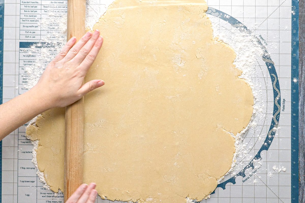 Overhead shot of hands rolling out dough with a rolling pin on a floured pastry mat marked with measurements