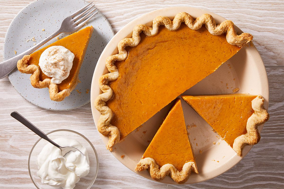 Overhead view of a classic Eggnog Pumpkin Pie with a slice removed, showcasing its creamy orange filling and fluted golden-brown crust; served with a spoonful of whipped cream and eggnog in a glass.