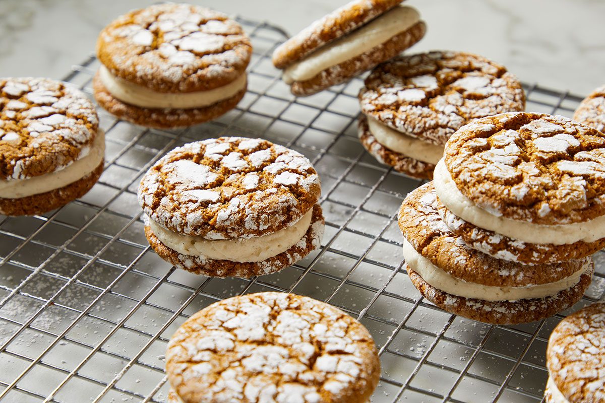 Close-up shot of Gingerbread Crinkle Cookie Sandwiches are arranged on a wire cooling rack over a marble background Each one has powdered sugar on top and creamy filling inside