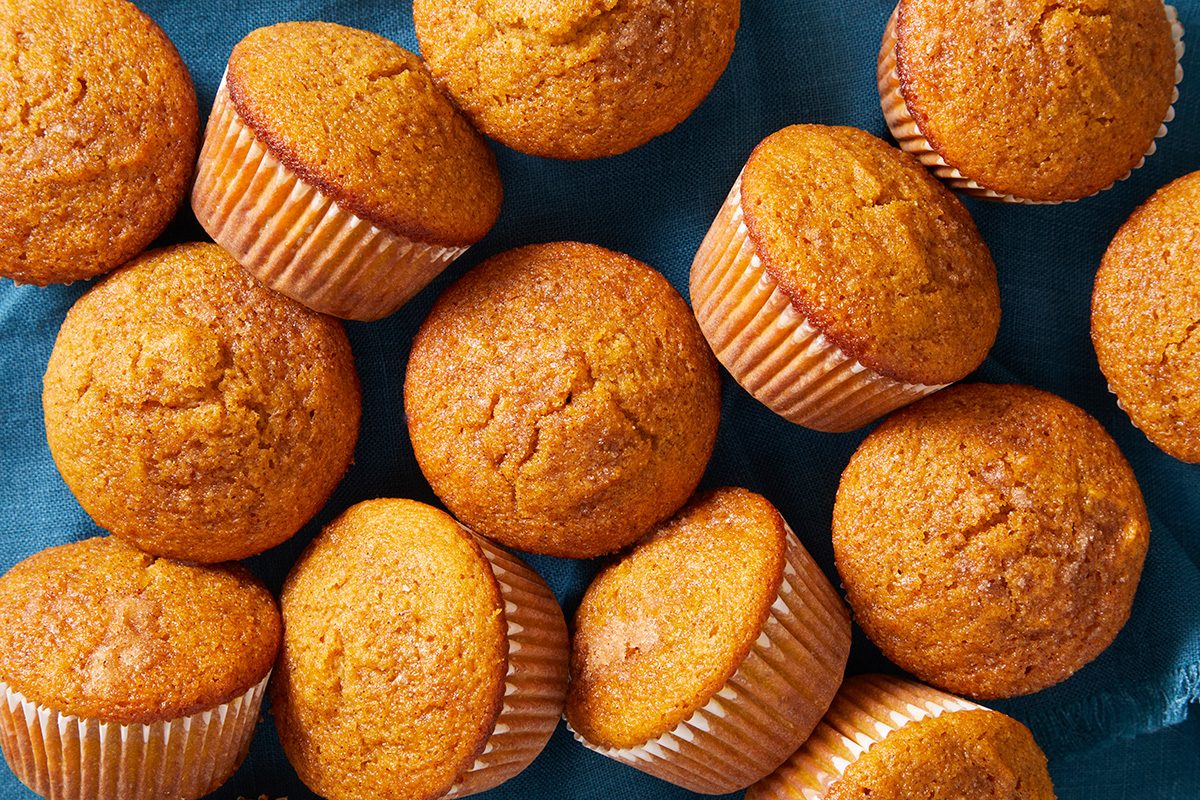 A group of golden-brown muffins in paper liners are arranged closely together on a blue surface.
