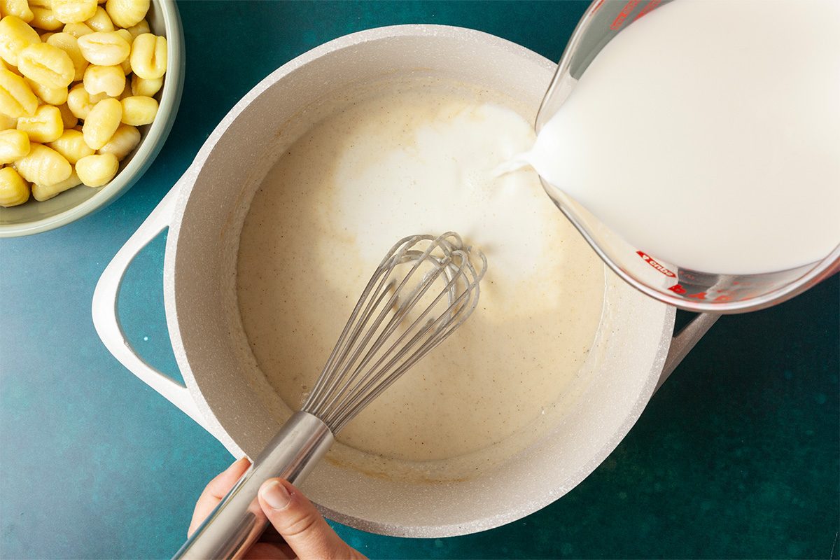 Overhead shot of a hand whisking a creamy sauce in a white pot as milk is poured from a measuring cup, with a bowl of uncooked gnocchi nearby on a green surface