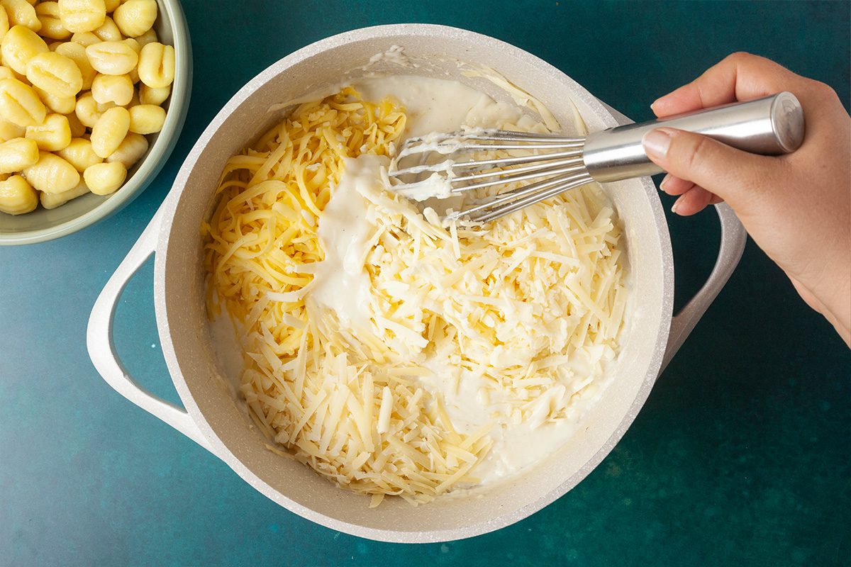 Overhead shot of a hand whisking shredded cheese into a creamy white sauce in a pot, with a bowl of uncooked gnocchi beside it on a teal surface