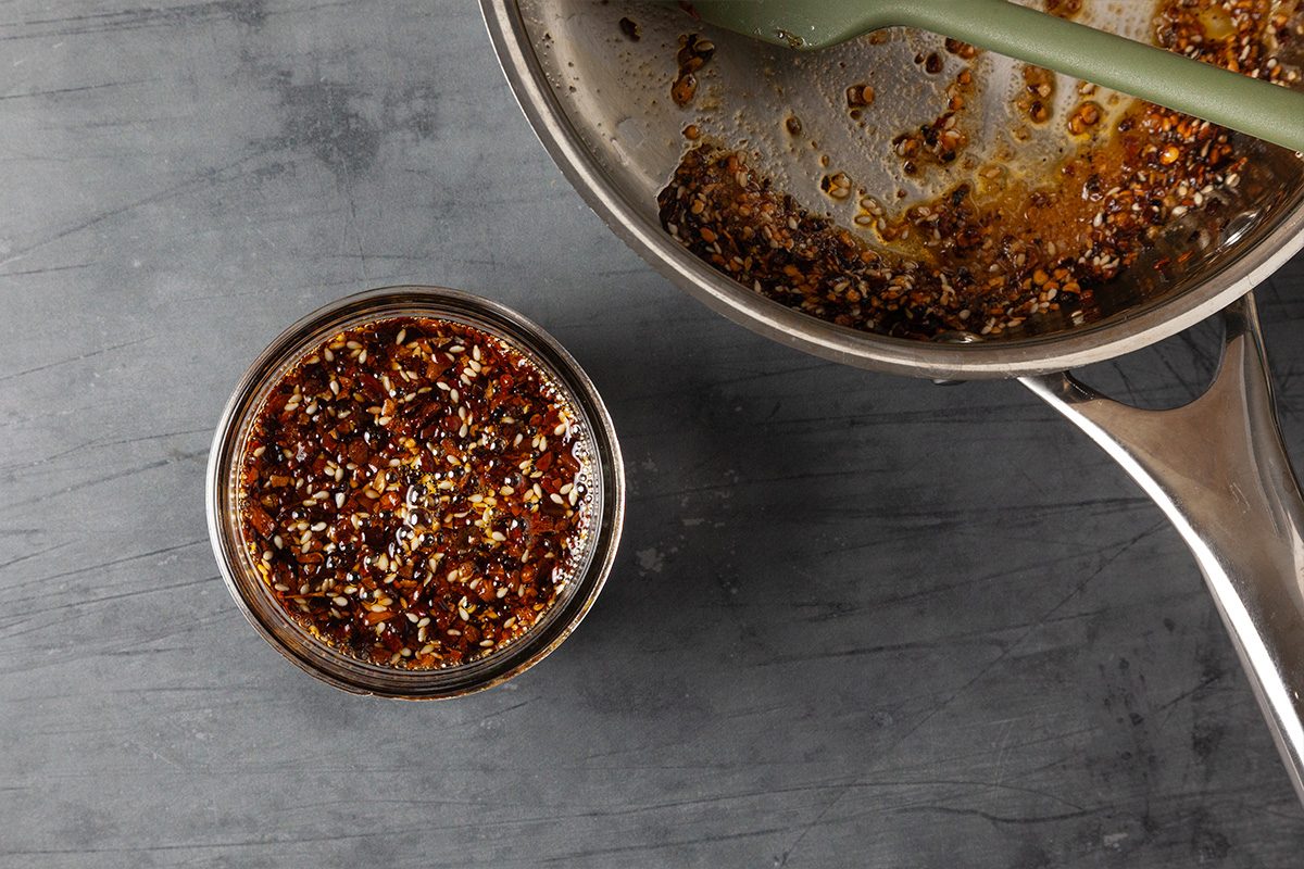 Overhead shot of freshly made chili crisp being poured from the saucepan into a small bowl, showing the rich, textured oil.