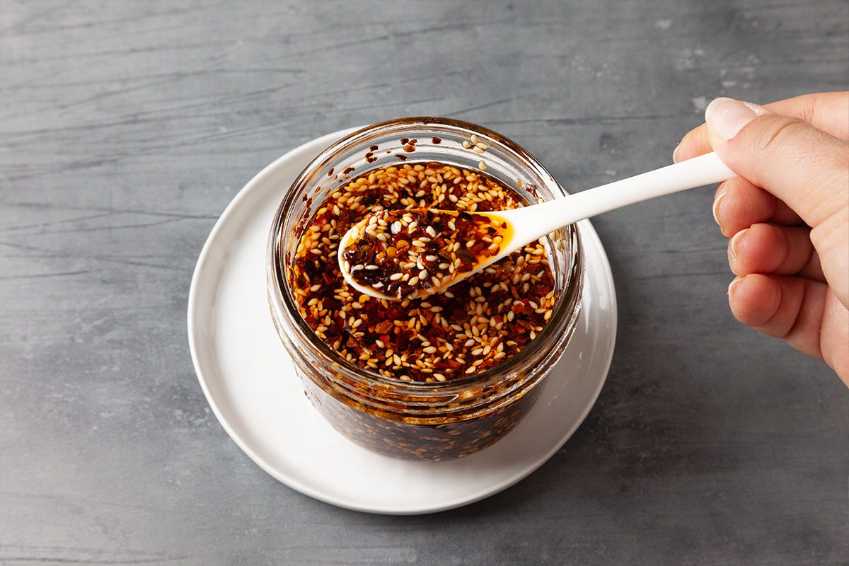 Close-up of a hand holding a spoonful of chili crisp above a small glass bowl, highlighting the crispy bits and deep red oil.
