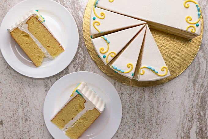 A round yellow cake with white frosting and colorful decorative swirls is sliced; two individual cake slices are served on white plates next to the remaining cake on a gold tray.
