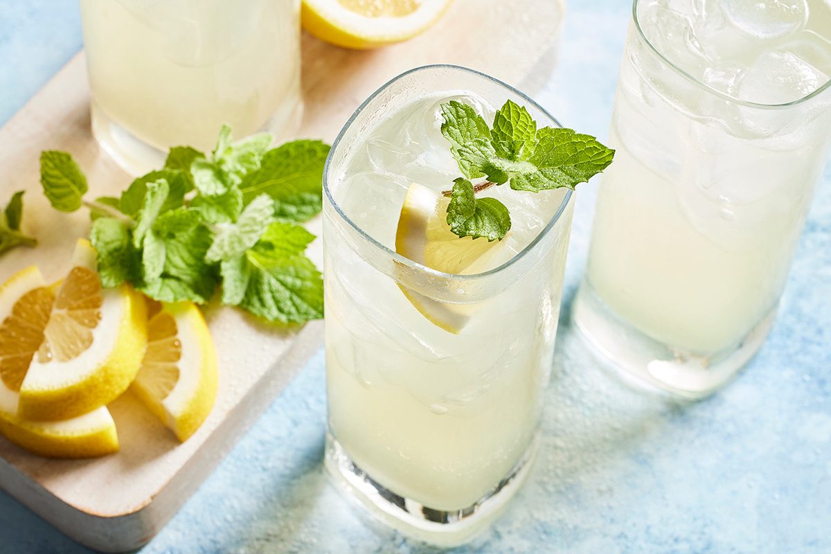 Three glasses of iced lemonade, garnished with lemon slices and mint sprigs, sit on a light blue surface. A small pile of lemon slices and fresh mint leaves rests on a wooden tray nearby.