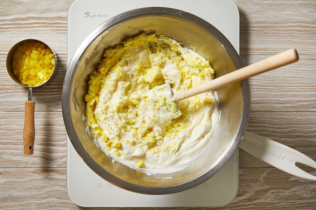 A metal mixing bowl filled with a yellow and white mixture is on a white surface, with a wooden spoon resting inside. Beside it, a measuring cup holds more diced yellow ingredients.