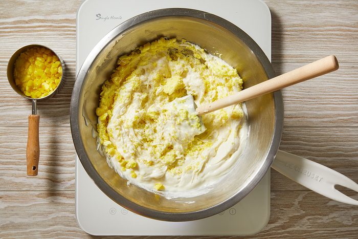 A metal mixing bowl filled with a yellow and white mixture is on a white surface, with a wooden spoon resting inside. Beside it, a measuring cup holds more diced yellow ingredients.
