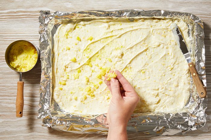 A hand sprinkles minced garlic over a rectangular sheet of dough spread on a foil-lined baking tray; a small cup with more garlic is on the side.