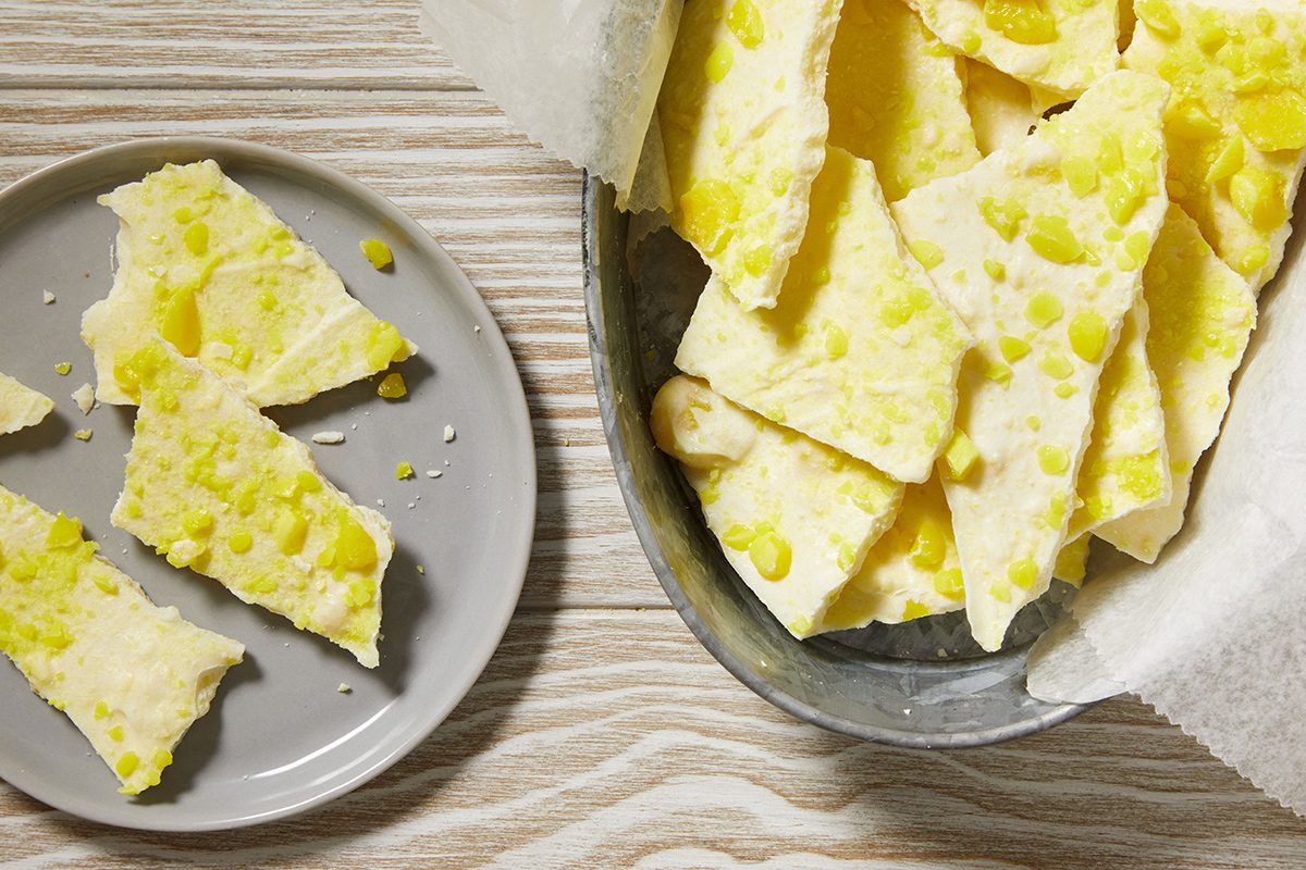 A metal bowl lined with parchment paper holds pieces of white chocolate bark topped with yellow candy bits. Nearby, a gray plate displays several broken pieces of the bark on a light wood surface.