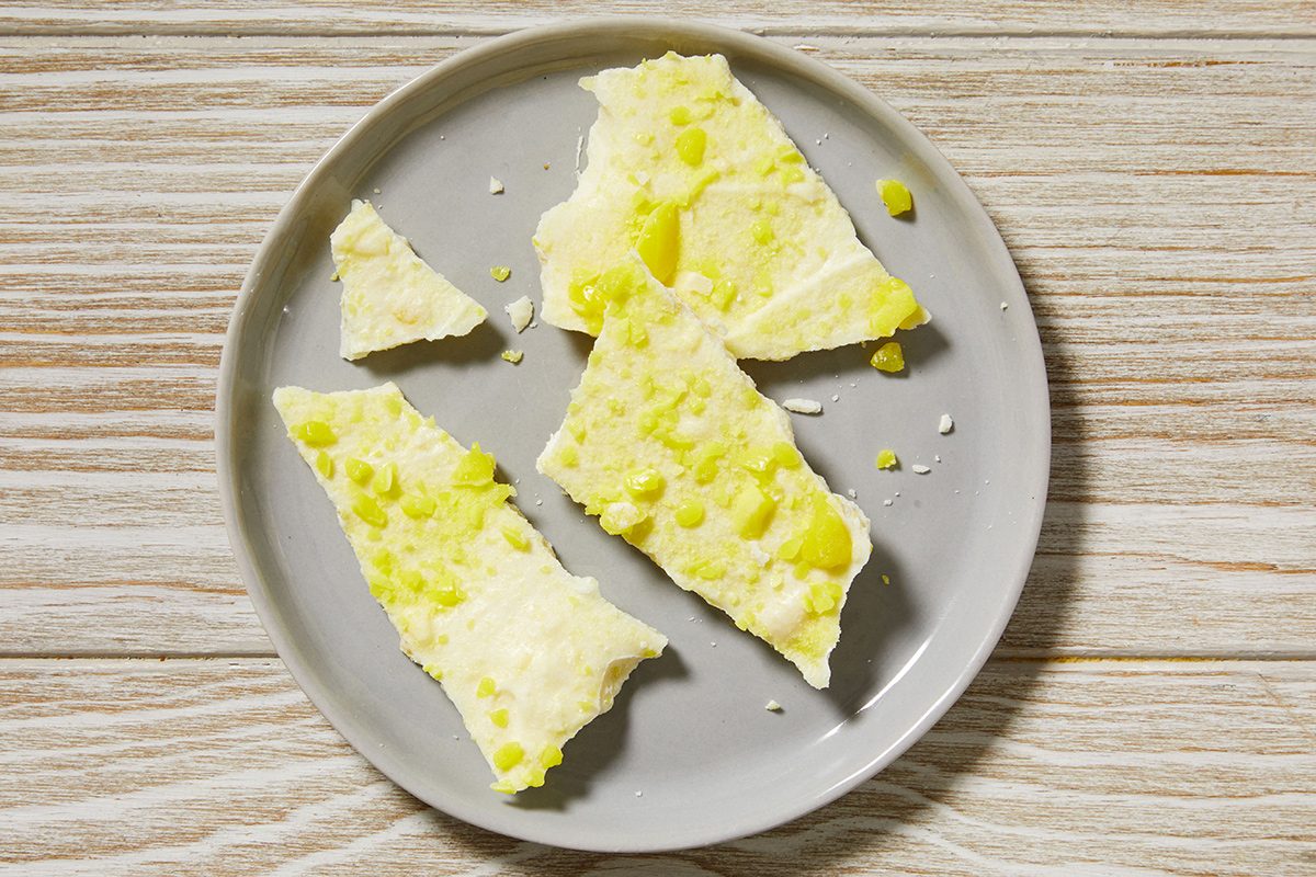 A grey plate holds several broken pieces of white chocolate bark topped with small droplets of yellow oil, set on a light wooden surface.