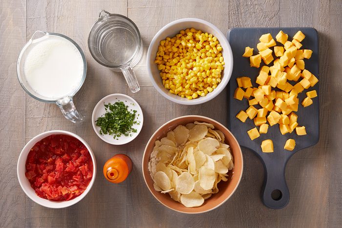 Ingredients on a table: a bowl of diced cheese, canned corn, diced tomatoes, chopped herbs, potato chips, a cup of milk, a cup of water, and an orange spice shaker.