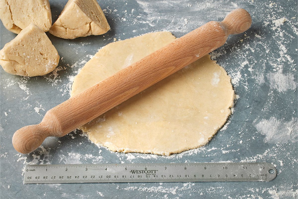 3/4 angle view of a wooden rolling pin resting on a round sheet of dough, with a ruler nearby on a floured surface and triangular dough pieces in the background;