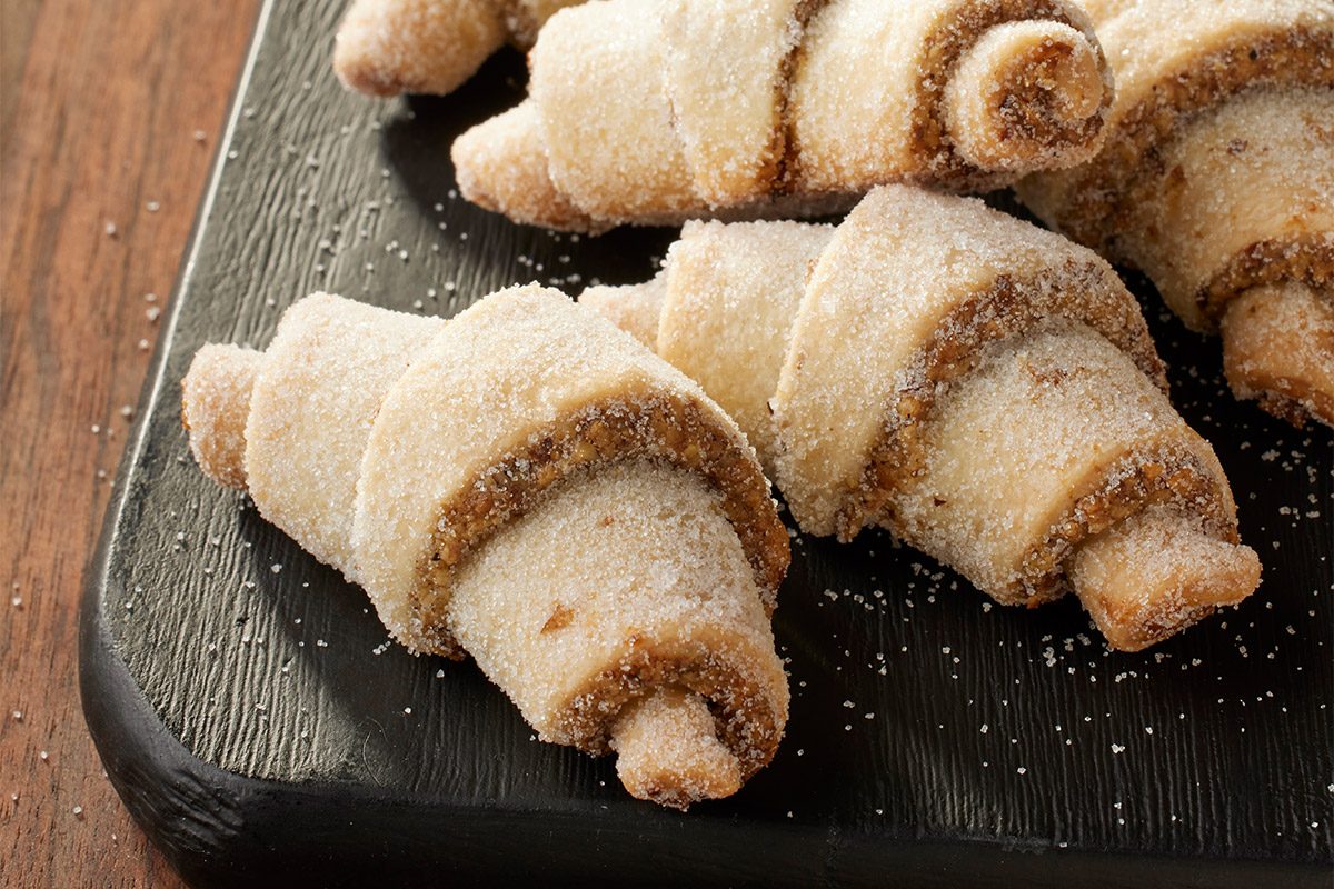 3/4 angle view shot of sugar-dusted Horn Walnut Cookies on a dark serving board, with additional cookies on a plate in the background, placed on a wooden table;