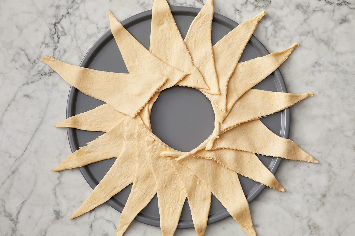 A ring of overlapping crescent dough triangles arranged in a sunburst pattern on a round baking sheet, placed on a marble surface.