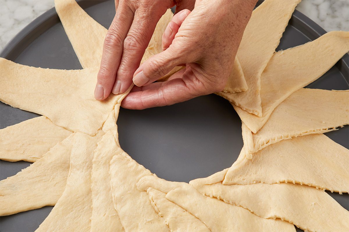 Close-up of hands arranging crescent roll dough triangles in a circular pattern on a baking tray, preparing a ring shape.