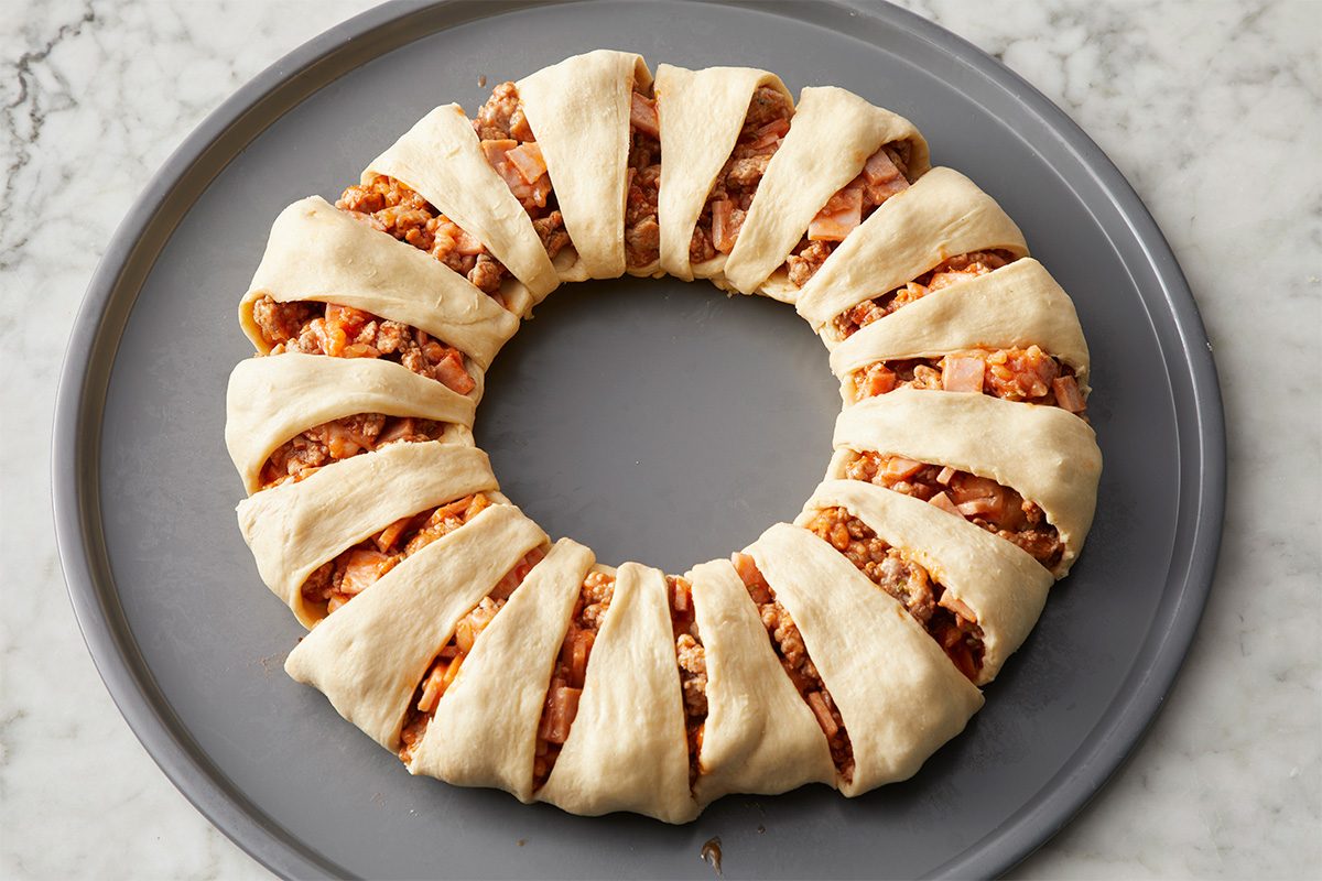 A round baking tray holds an unbaked crescent ring filled with a chunky meat and cheese mixture. The dough is sliced and folded over the filling, creating a wreath shape. The tray sits on a marble countertop.