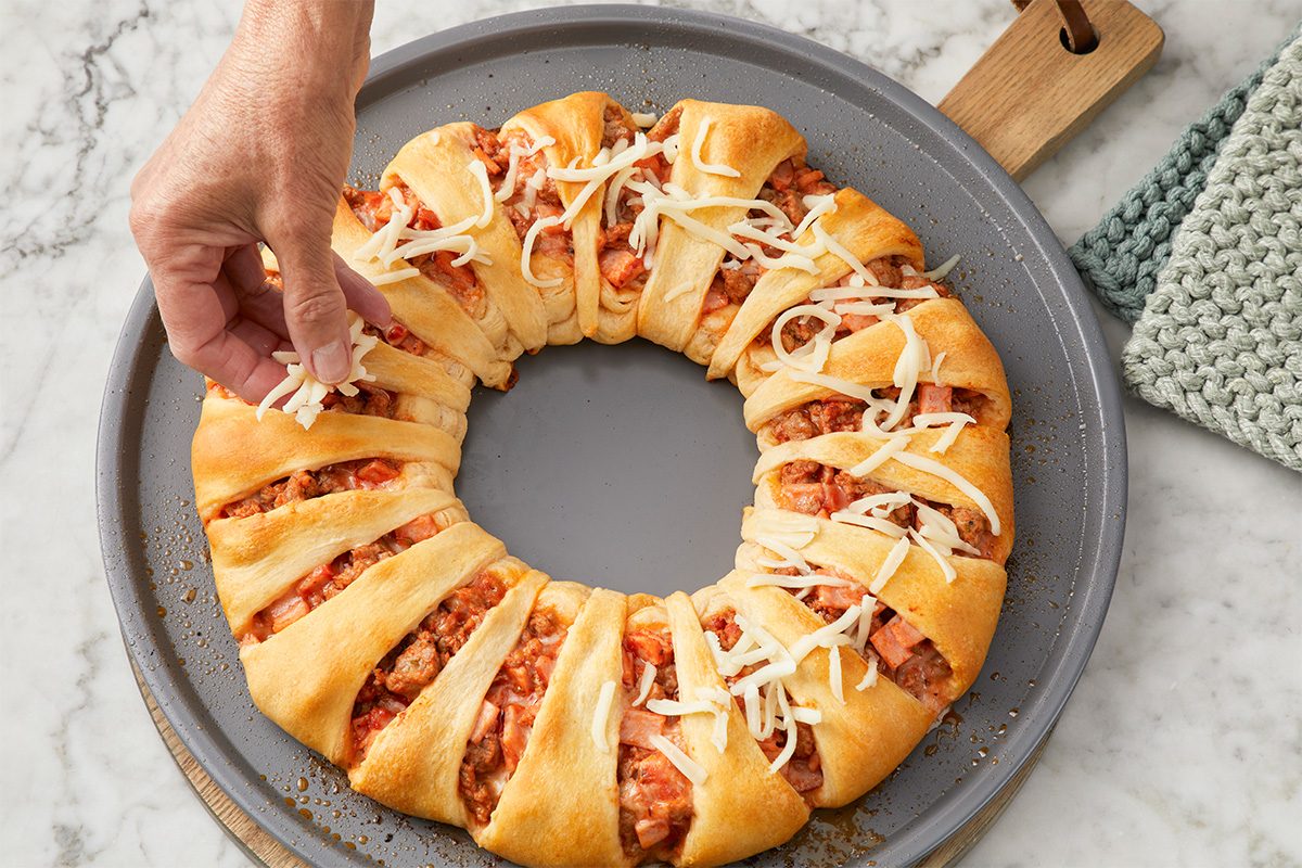 A hand sprinkles shredded cheese onto a baked crescent ring filled with a meat and tomato mixture, presented on a round baking tray atop a marble surface.