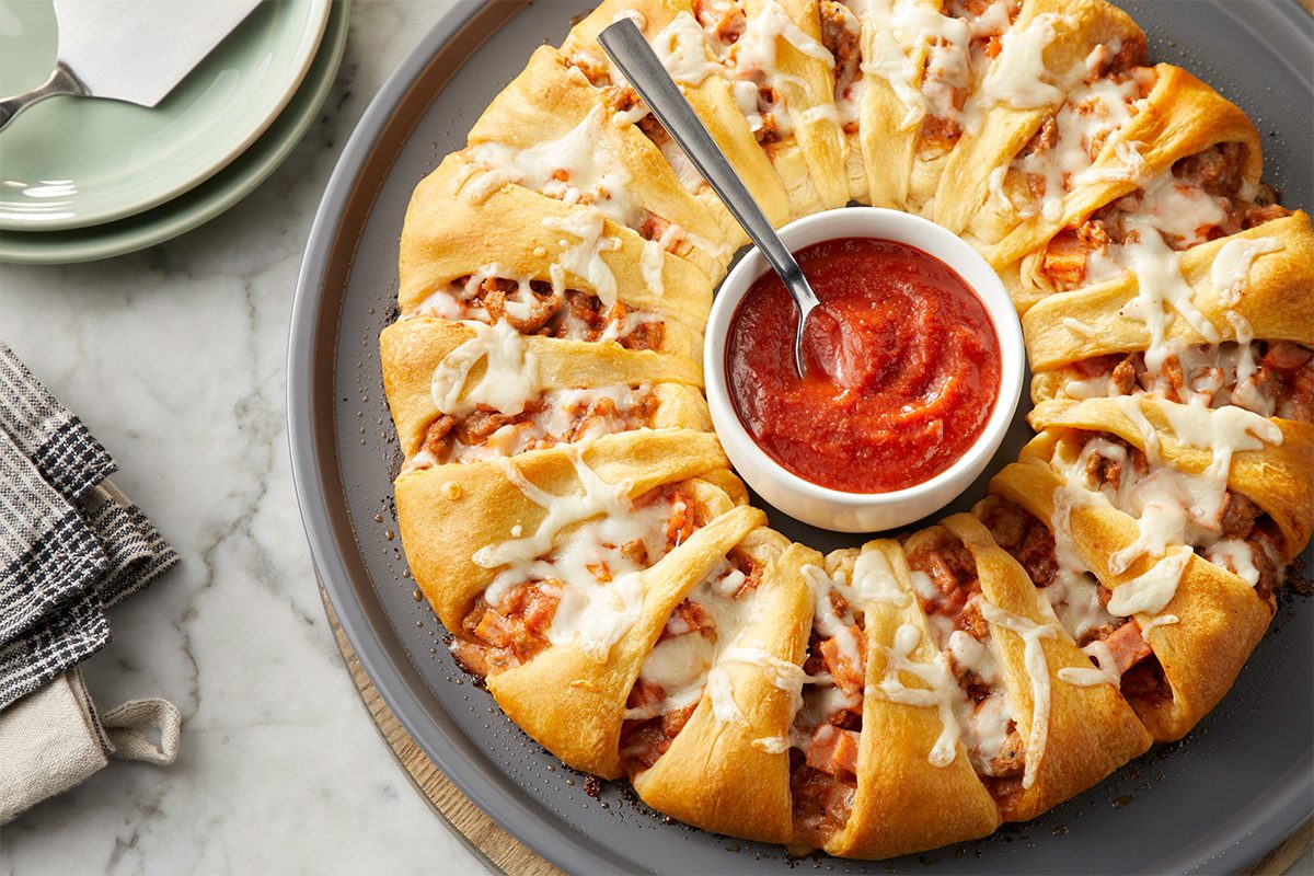 A round tray holds a ring of baked crescent rolls stuffed with cheese and filling, topped with melted cheese. A small bowl of marinara sauce sits in the center with a spoon for dipping. Plates and napkins are nearby.