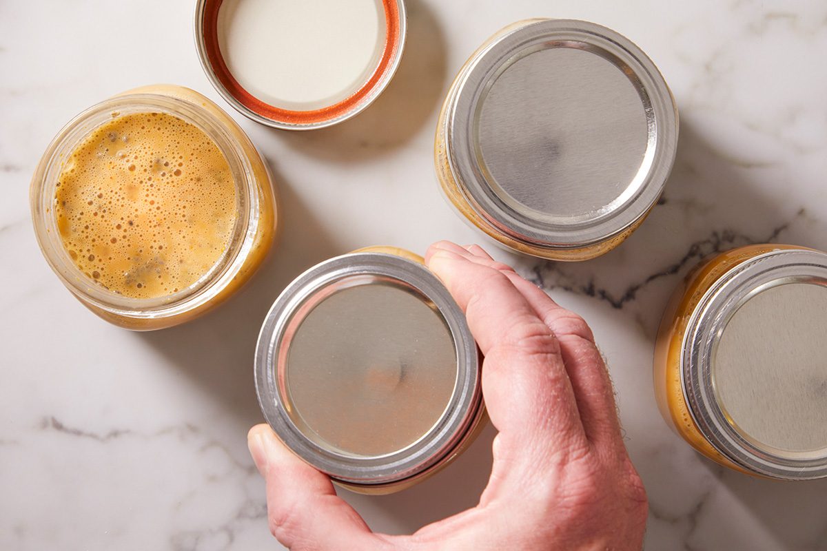A hand is holding a glass jar with a silver lid, surrounded by three similar jars on a marble surface. One jar has its lid off, revealing a frothy, orange mixture inside.