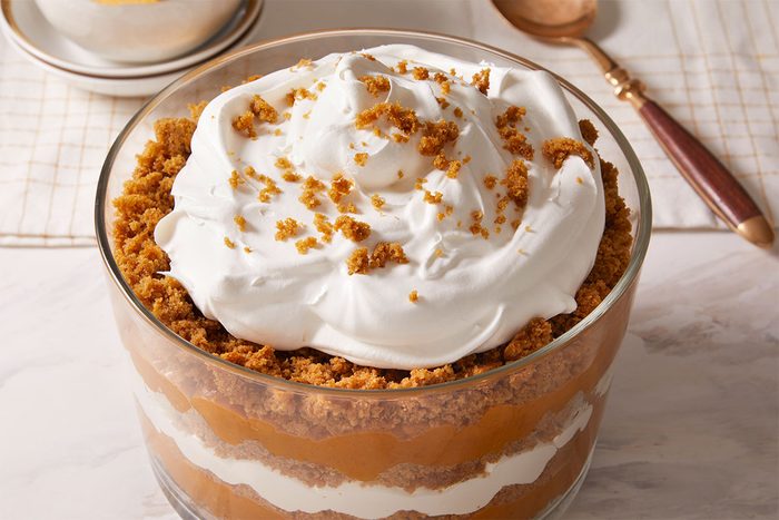A layered dessert in a glass bowl, featuring alternating layers of whipped cream and a brown crumb mixture, topped with whipped cream and sprinkled crumbs. A spoon and a bowl are in the background on a marble surface.