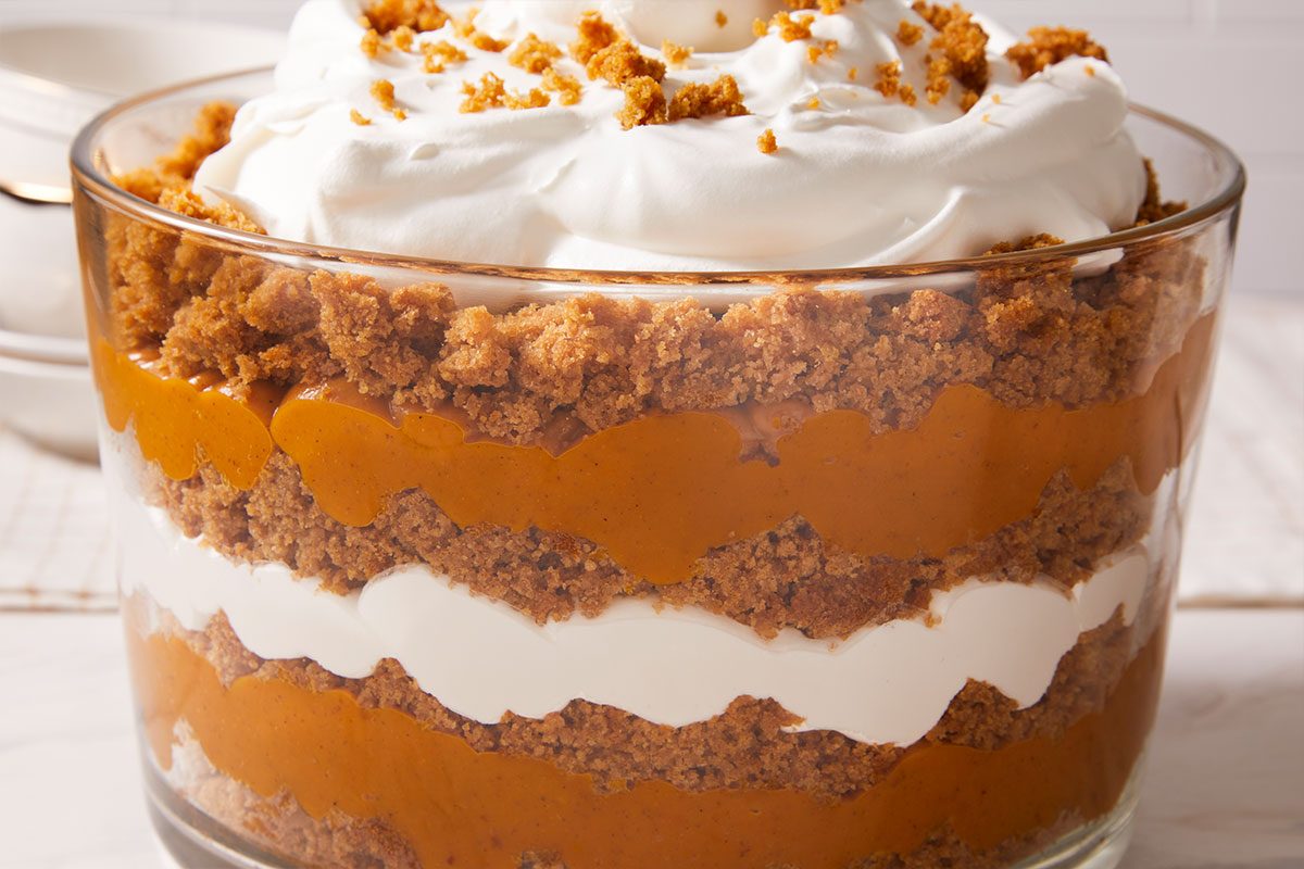 A glass trifle dish filled with alternating layers of whipped cream, pumpkin filling, and crumbled cookies or cake, topped with more whipped cream and cookie crumbs.