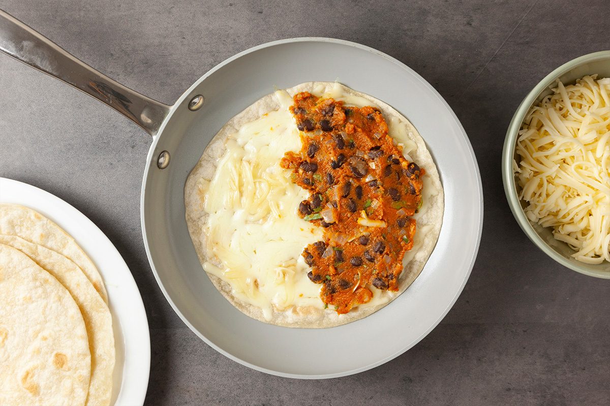 Overhead shot of a skillet with a tortilla topped with melted cheese and a black bean–tomato mixture, set on a gray surface beside a bowl of shredded cheese and a plate of plain tortillas;