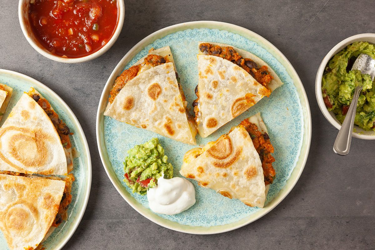 Overhead shot of two plates, each with three Pumpkin Quesadilla wedges served with sour cream and guacamole; bowls of salsa and extra guacamole sit nearby;