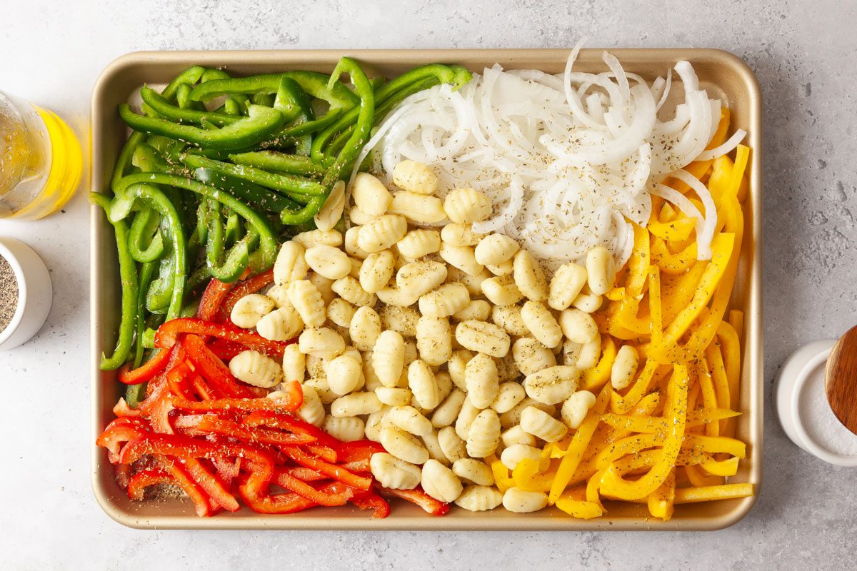 Overhead ingredient shot of a baking tray filled with rows of uncooked gnocchi, ringed by sliced tri-color bell peppers and onions, all dusted with black pepper.