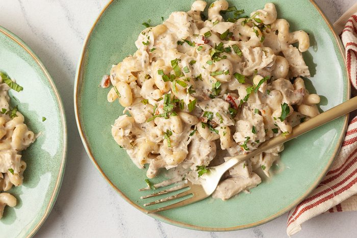 Overhead shot of a green plate of creamy Slow-Cooker Chicken Boursin Pasta mixed with chicken and garnished with chopped herbs, with a fork on the side and a striped napkin nearby