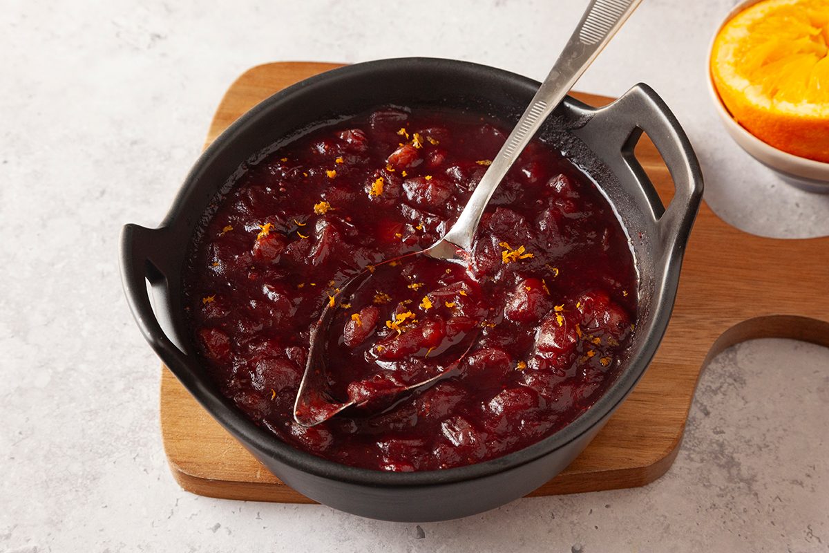 A black bowl filled with chunky cranberry sauce sits on a wooden board. A metal spoon rests in the bowl, and a half orange is visible in the top right corner.