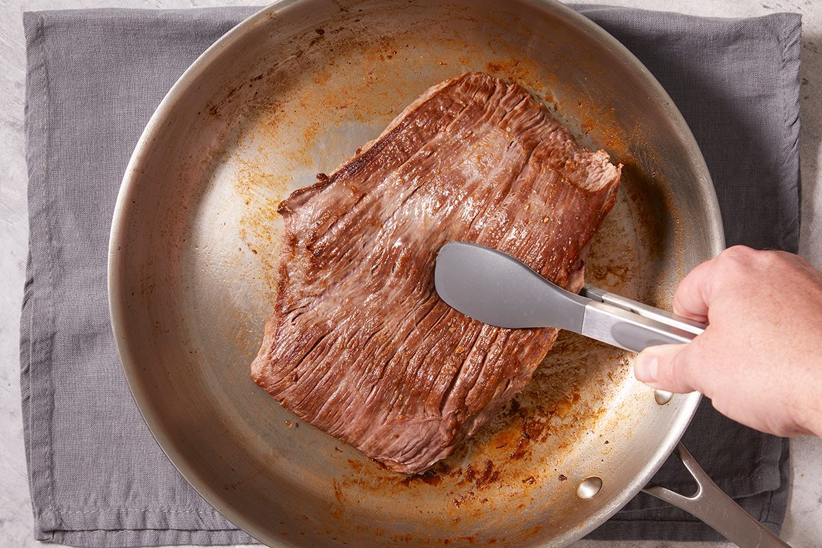 A hand uses metal tongs to sear a large piece of beef in a stainless steel skillet, which is set on a gray cloth.