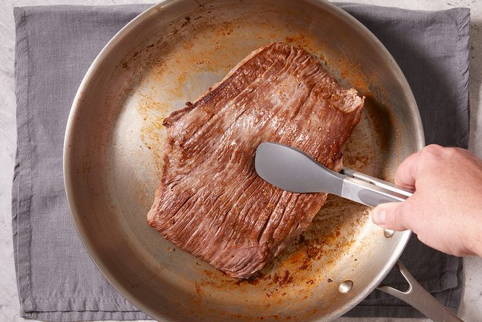 A hand uses metal tongs to sear a large piece of beef in a stainless steel skillet, which is set on a gray cloth.