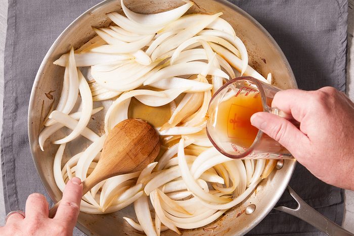 Sliced onions are being sautéed in a skillet; a person stirs with a wooden spoon while pouring liquid from a measuring cup over the onions.