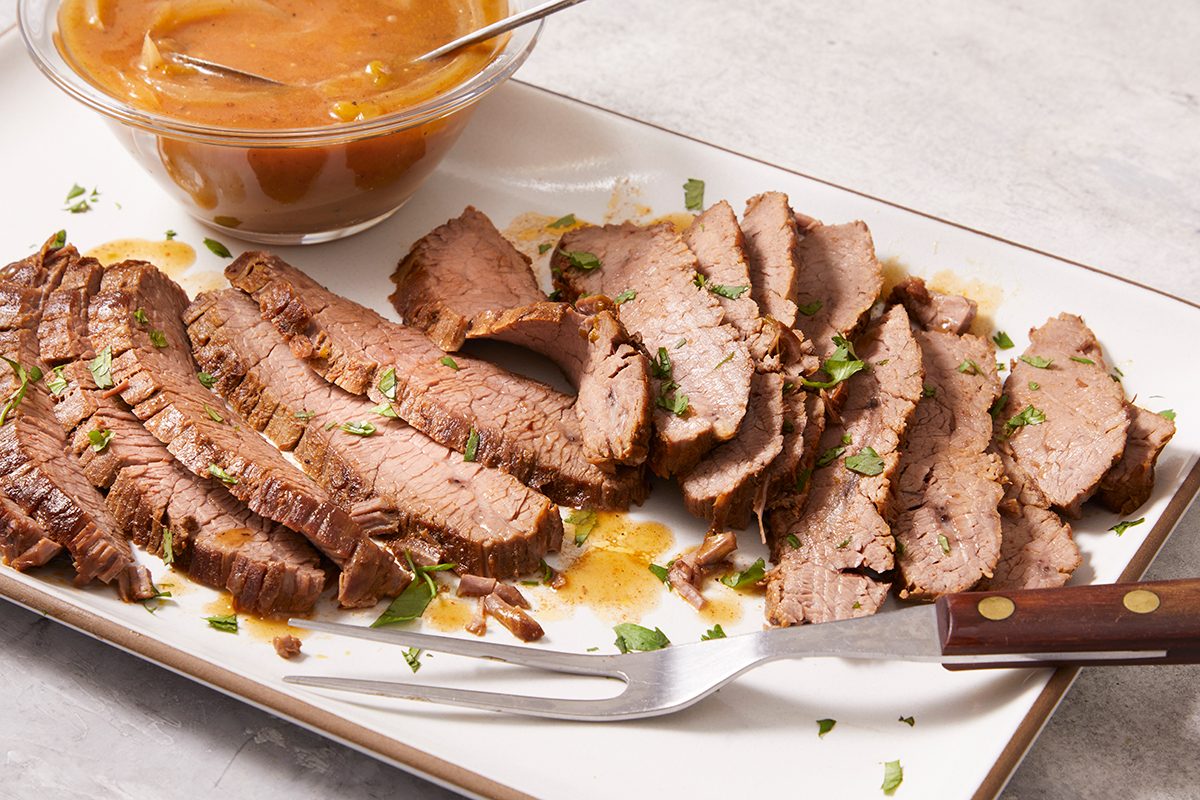 A platter of sliced cooked beef garnished with herbs is served with a brown sauce in a small bowl and a serving fork placed on the side.