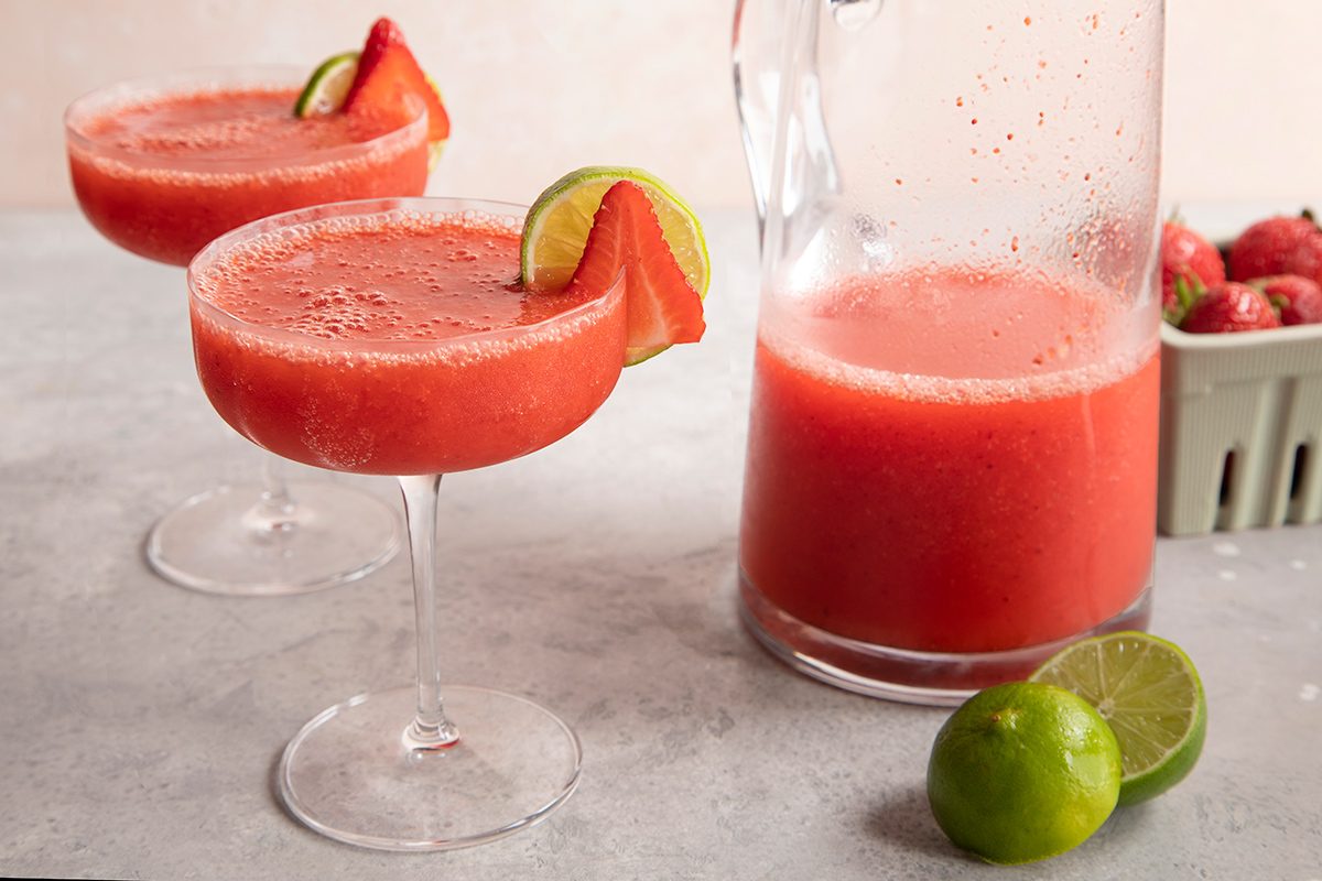 Two glasses of strawberry margarita garnished with lime and strawberry slices, next to a pitcher of the drink, fresh limes, and a container of strawberries on a light surface.