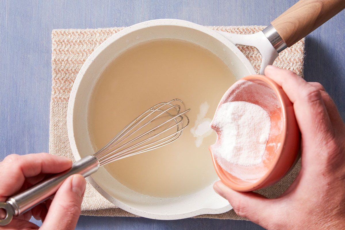 A person holds a whisk in a bowl of liquid while pouring a white powder from a small bowl, preparing ingredients on a light-colored surface with a textured cloth underneath.